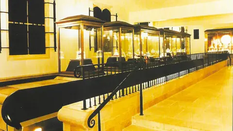 Paul Dennis The Jewel Room at the Tower of London. Five glass-sided display cases containing various crowns and other items of the Crown Jewels sit on a platform. In front of them are wrought iron railings designed to separate visitors from the cases as they file past. The room is brightly lit, with stone flooring.