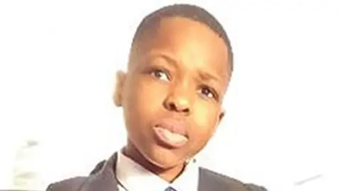 Daniel Anjorin wearing school uniform of blazer, v-necked jumper, shirt and tie. He is looking toward the camera and is sitting on the back seat of a bus.