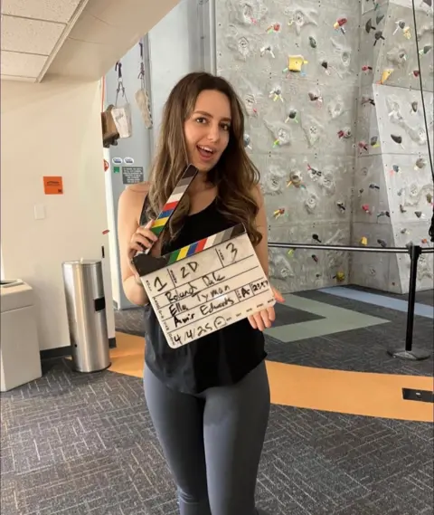Ella Tyman Ella holds a director slate, posing for the camera. Behind her is a climbing frame. She is inside. 