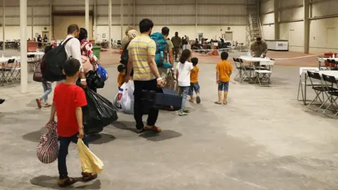 Getty Images Afghan Special Immigrants including women and children carrying bags walk through the in-processing building which is a large warehouse on August 20, 2021 at Camp As Sayliyah, Qatar. Soldiers help process Special Immigration Applicants seeking relocation to the United States. 