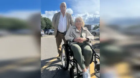 Nick and Hilary Saul in the car park. Mr Saul stands behind Mrs Saul, who is sitting in a wheelchair