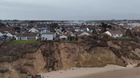 A drone image of a sandy cliff edge, with a line of chalet properties just a few feet from the cliff edge.