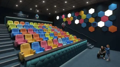 Manchester Foundation Trust Charity A CGI image shows five rows of colourful cinema seats with space at the front for wheelchairs or hospital beds. A woman and child are looking at the cinema while a man and child are standing near the top of a stairway at one side of the row of seats. A sign at the back says 'MediCinema Feel better with film'.
