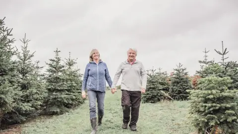 Zoe Harris Photography Alastair and Diane stood arm in arm in a field. There are Christmas trees around them. 
