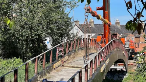 A bridge covered in chains and grass as seen from one end. It's being lifted by an orange crane backed by trees and blue skies.