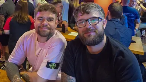 Sam Hulme Two men, both with dark hair and beards, smile as they sit at a table inside a crowded bar.