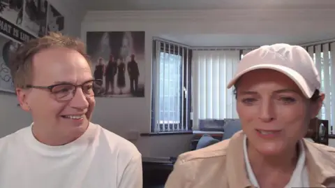 Andy Reiss and Katie Leeming. Andy wears black-rimmed glasses, has short blonde hair and wears a white T-shirt. Katie has a pink baseball cap on and wears a beige jacketed over a white top. They sit in a room with blinds on the windows.