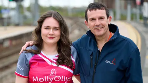 Two people standing smiling. A woman with long brown curly hair wearing a bright pink top which says Molly Ollys on it. The man has short brown hair and is also smiling. He has his arm around his daughter. He is wearing a navy jacket with avanti west coast on it. 