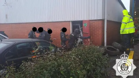 West Mercia Police Five police officers in black tactical clothing a lined up in single file as they enter an industrial unit with their faces obscured by black dots added to the photo. A police officer wearing a yellow high viz coat watches on from the side.