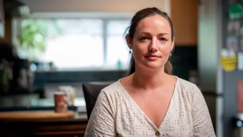 A woman wearing a white top sits in her kitchen with cups and a window in the background