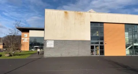 A modern school building in red brick and beige stone with large two-storey windows and the Auchmuty High school crest on the wall. The playground outside is tarmac-ed.