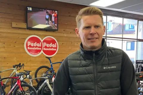 Clare Worden/BBC Matthew Watts stands in front of a row of bikes and a red sign reading PEDAL REVOLUTION. He has blond hair and is wearing a black puffer jacket. 