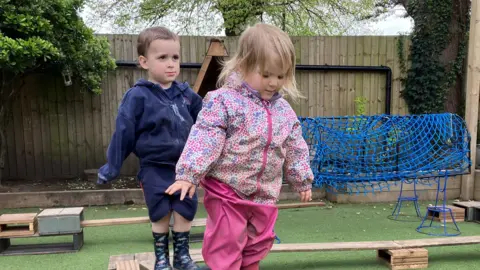 Two toddlers are walking on a wooden walkway made of planks and steps in a nursey garden setting. The boy at the rear has short light brown hair and is wearing a navy blue fleece and navy shorts with navy wellies with dinosaur pattern, and the girl in front has straight blonde hair and is wearing a pink and blue flowery raincoat, pink waterproof trousers and pink wellies. More walkways, a few inches off the ground are visible in the background, as well as a tunnel made of blue netting. 