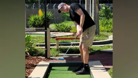 James Goatley A man in a black shirt and beige shorts putting on a mini golf course.