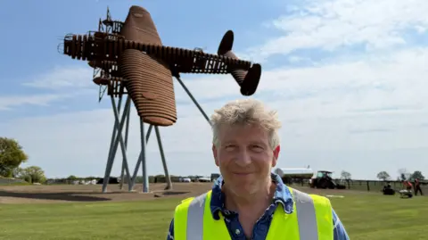 Charlie White has grey hair and a blue shirt. He is also wearing a yellow hi-vis on top. Behind him is a large steel brown sculpture of a Lancaster bomber in a flying motion and tilted to the side.