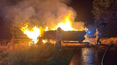 Peterchurch Fire Station Two firefighters use garden forks to tackle a trailer fire. The trailer is carrying hay bales which are well alight. 