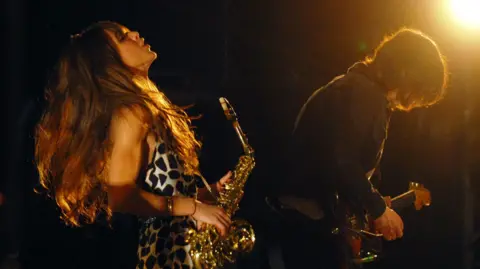 Abi Harding, wearing a black and white spotted dress and holding a gold saxophone, throws her long dark hair back, as Dave McCabe, with long dark hair and a black top, leans over his guitar in performance