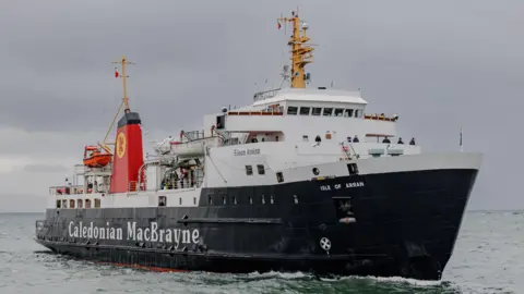A Caledonian MacBrayne ferry sailing at sea under a grey sky, with passengers visible on deck and the ship’s name and Isle of Arran marked on the bow.