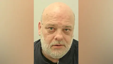 Anthony Kingston's mugshot. He has a bald head, a short white beard and is wearing a black top against a pale grey background.