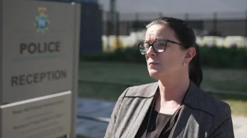 Det Ch Supt Karen Jaundrill, who has black hair pulled into a ponytail, black rimmed glasses and wearing a grey blazer over a black top, speaks to the camera outside a grey sign bearing the Merseyside Police insignia