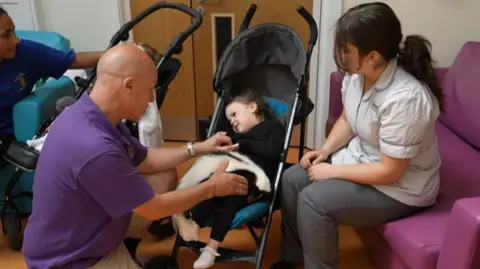 A young girl with brown hair sits in a pram with a skunk on her lap. A nurse in scrubs is to her right and a man in a purple shirt to her left.