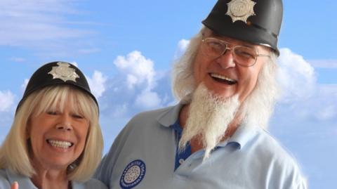 Clive, with white beard, stood with his wife, Jackie, both wearing blue shirts in front of a cloudy blue sky. They are both wearing traditional style police helmets. 