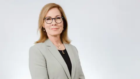 A woman wearing glasses is smiling at the camera. She is posing for a professional headshot. She has shoulder length blonde hair, a grey blazer and black top.