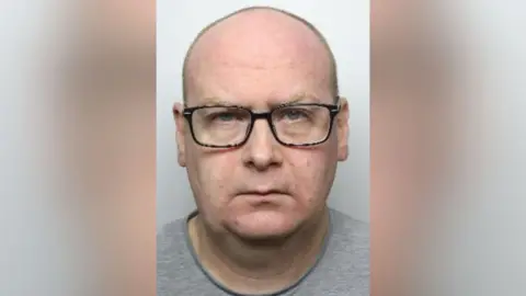 Police mugshot of Lee Maidens, a bald man with ginger/blond eyebrows and square glasses.