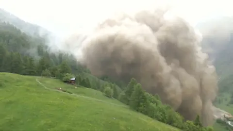 A large cloud generated by rock and mud rises over the green slope of a valley