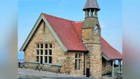 Supplied Cullercoats Watch House, a beige stone-brick building with an arched roof of red tiles. The roof is lined by woodwork which is painted green. A similarly painted fence surrounds the building where it looks onto the sea, underneath a canopy where the roof juts out. The most striking feature is that it has a clock tower with a spire which rises into the sky.
