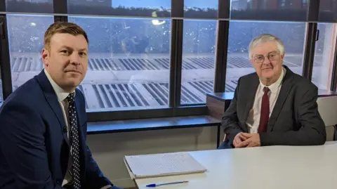 The image shows Mark Drakeford and BBC correspondent Cemlyn Davies sat at a table. Drakeford is wearing a grey suit, white shirt and red tie. Davies is wearing a navy suit, white shirt, and blue and white spotty tie. Both are looking at the camera.