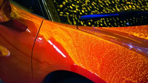 A car, showing part of its front, a door, the colour looks orange with reflections on it. 