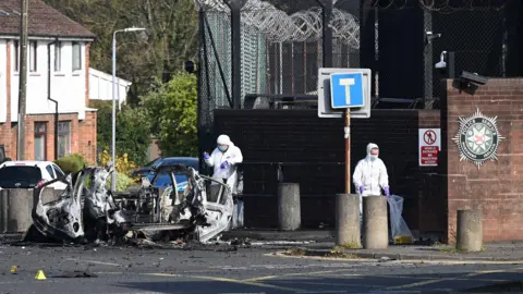 Charles McQuillan/Getty Images Two forensic investigators at the scene of the Dunmurry car explosion. They are both wearing white hazmat suits, blue masks and purple plastic gloves. They are walking around the wreckage of the exploded car which is lying on the road beside concrete bollards outside the large police station wall. On investigator is carrying a yellow evidence marker inside a clear plastic bag. 