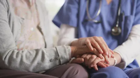 Two people sit side by side with their hands placed one on top of the other. Only their hands, arms and torsos are visible, with the person on the right in a blue nurse's uniform and wearing a stethoscope.