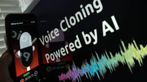 This picture is has a touchscreen mobile phone being held next to a screen which reads "voice cloning powered by AI". The mobile phone screen display has a animated radio microphone with a red background. The lower third of the mobile phone screen has a record button and a timing display. 