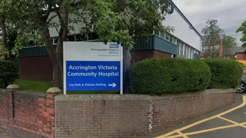 Google A sign outside the Accrington Victoria Community Hospital. It has directions to the car park and visitor parking. The sign is next to trees and bushes and surrounded by a brick wall.