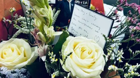 Stevie Moore A close up of a bunch of flowers with white roses and bunches of leaves. There is a handwritten note in the bunch.