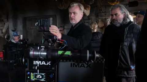 Universal Pictures Christopher Nolan in the centre of the photo with cinematographer Hoyte van Hoytemaon on the right. Nolan is looking through Imax camera. Both are wearing dark clothes.