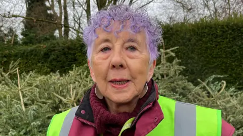 Tom Jackson/BBC Sheila Pierre is in the middle. She has purple short curled hair and is wearing a burgundy roll neck jumper and a yellow Hi-Vis jacket.