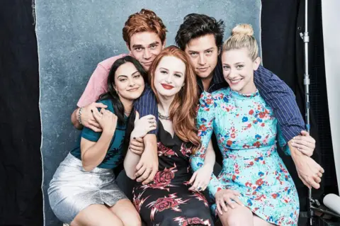 Getty Images Kj Apa, Cole Sprouse, Camila Mendes, Madelaine Petsch and Lili Reinhart pose for a photo together in front of a paper background like those often seen in school photos. The two male cast members stand behind the three female co-stars, with their arms around them, leaning in close. They look relaxed and comfortable in each others' company. 