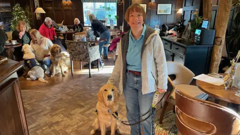 BBC/Jack Hadaway-Weller A woman in a blue polo shirt and a grey jacket stands with a golden retriever dog in a busy pub.

In the background are several other dogs and people having drinks.