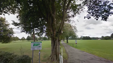 Google A wide view of Queens Park in Caterham, Surrey, showing trees in full leaf and lawns with a concrete path running between them.