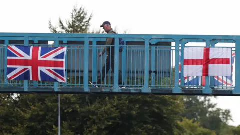 Reuters A man wearing a cap and rucksack is walking across a blue bridge over a main road. On the side of the bridge are a Union Flag and the St George's flag.