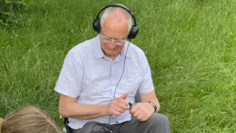 A man sat on a chair in the grass. He has on headphones which are connected to a small morse key in his hands with a black wire. He is wearing a light blue shirt with white stripes and grey canvas trousers. He is also wearing some clear rimmed glasses.