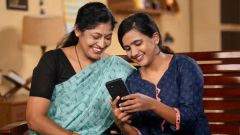 Getty Images A representative image of two Indian women smiling and looking at a mobile phone.