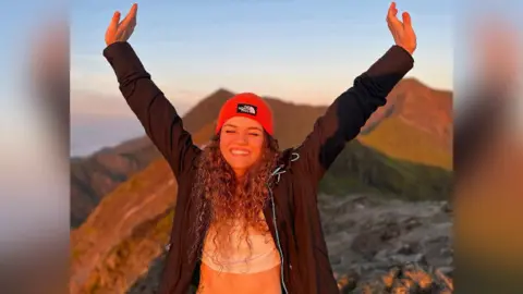 Jamie Graham A picture of Maria Eftimova smiling with her eyes shut and her hands in the air. There are mountains behind her and the picture is taken in a sunset. Maria has curly hair with a red hat on. 