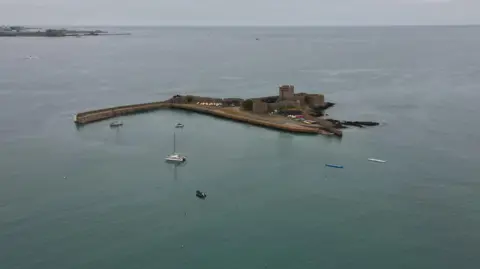 A drone shot of St Aubin's Fort. It is a concrete structure in the sea, with several small boats surrounding it.