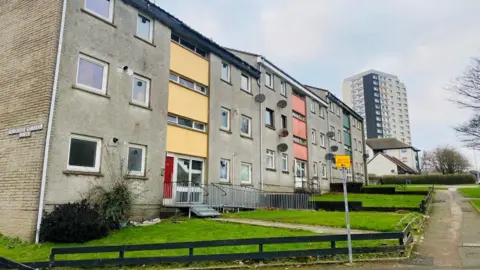 Blocks of flats, both three-storey and a high rise, with green gardens, next to a road, under a cloudy blue sky.