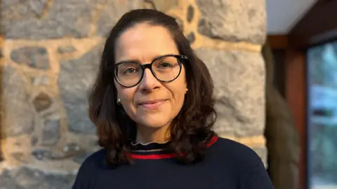 BBC Deputy Andrea Dudley-Owen is smiling at the camera as she stands in front of a stone wall. She has shoulder-length dark hair pinned back from her face and is wearing a pair of glasses.