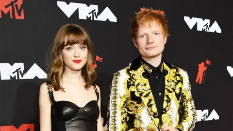 Getty Images Maisie Peters and Ed Sheeran on the red carpet at an MTV event. A young Maisie Peters has brown hair and wears black ankle boots with heels and a black leather dress with buckle straps. Ed Sheeran wears a yellow paisley patterned blazer with black trousers and shoes and a black shirt underneath